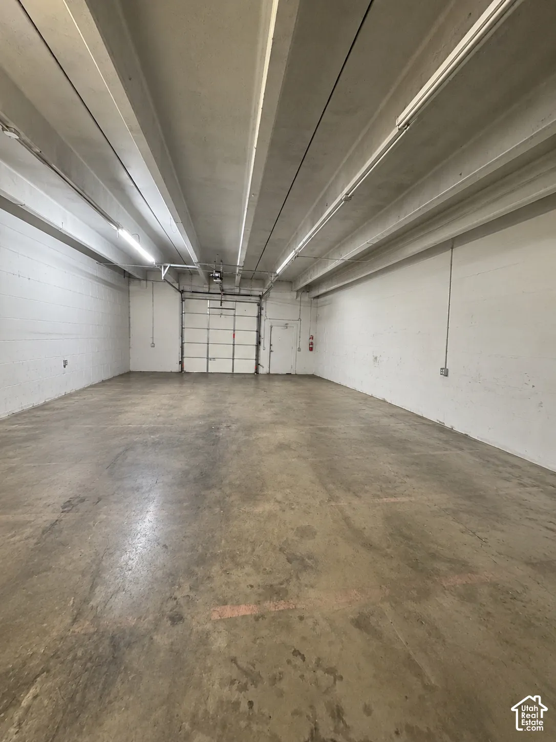 Spacious interior featuring exposed concrete walls, high ceilings with exposed structural beams, industrial concrete flooring, and a large metal roll-up door
