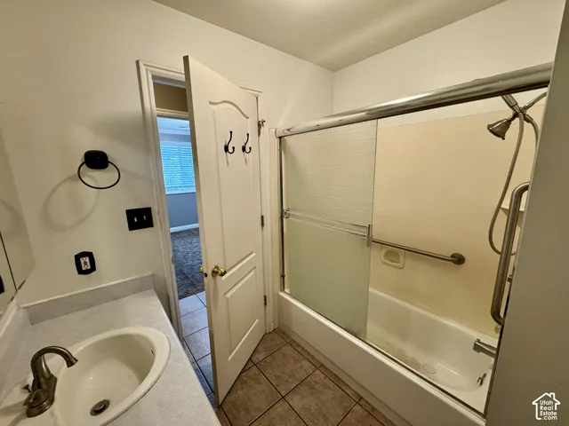 Bathroom featuring a single-basin vanity with a solid-surface countertop, tile flooring, and a bathtub with a sliding glass enclosure and wall-mounted grab bar