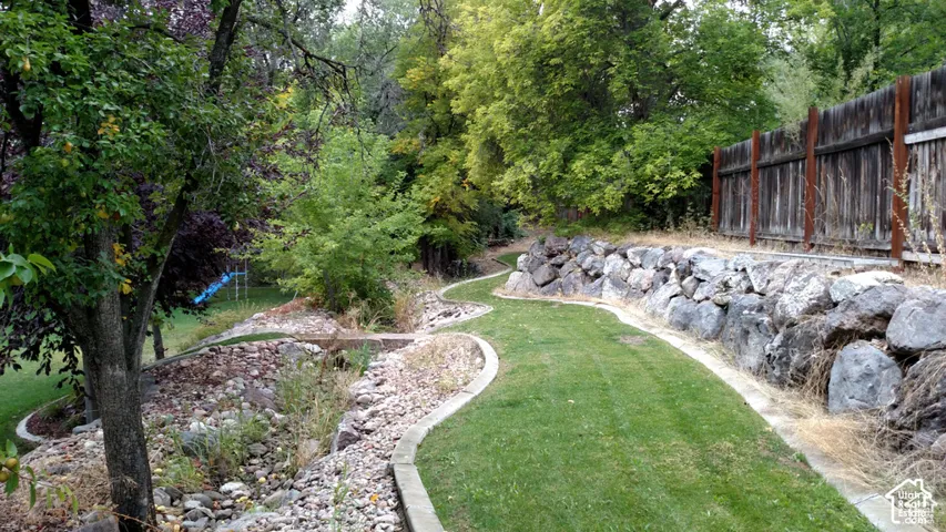 Backyard featuring tiered stone retaining walls, mature trees, manicured lawn, stone landscape beds, and wood fencing