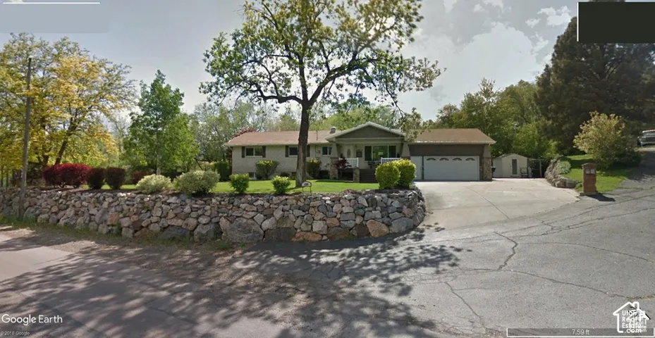 Exterior featuring stacked stone retaining wall, concrete driveway, attached multi-car garage, mature landscaping, and exterior lighting