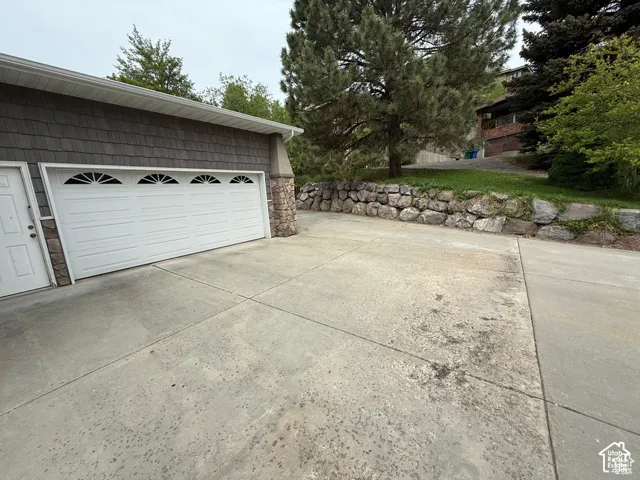 Multi-car garage featuring decorative sunburst window detailing, stone corner accents, shingled siding, and an expansive concrete driveway bounded by a stacked stone retaining wall