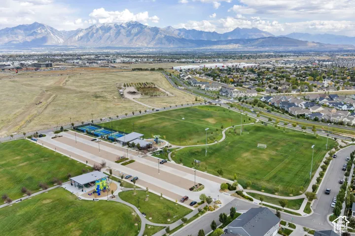 Aerial perspective of a community park featuring tennis courts, paved multi-use paths, expansive grassy fields, playground equipment, and mountain range scenery