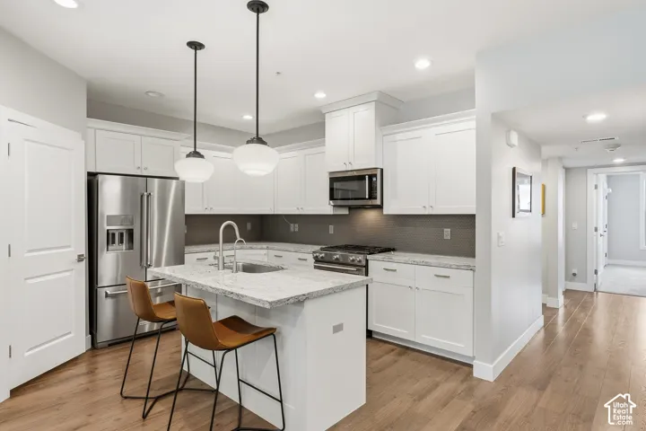 Kitchen featuring white cabinetry, stone countertops, stainless steel appliances, a central island with a built-in sink, and wood-finish flooring