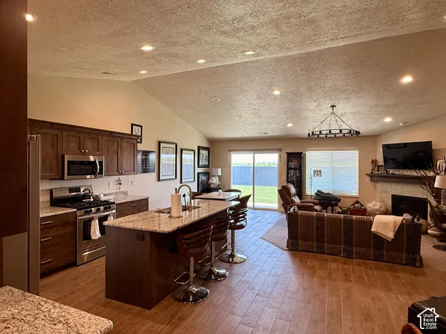 Open-concept area featuring vaulted ceilings, recessed lighting, wood-finish flooring, a kitchen island with stone-style countertops, and a fireplace with a stone-style hearth