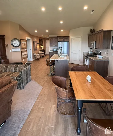 Open-concept kitchen featuring stained wood cabinetry, stone countertops, a central island with sink, recessed lighting, and wood-finish flooring
