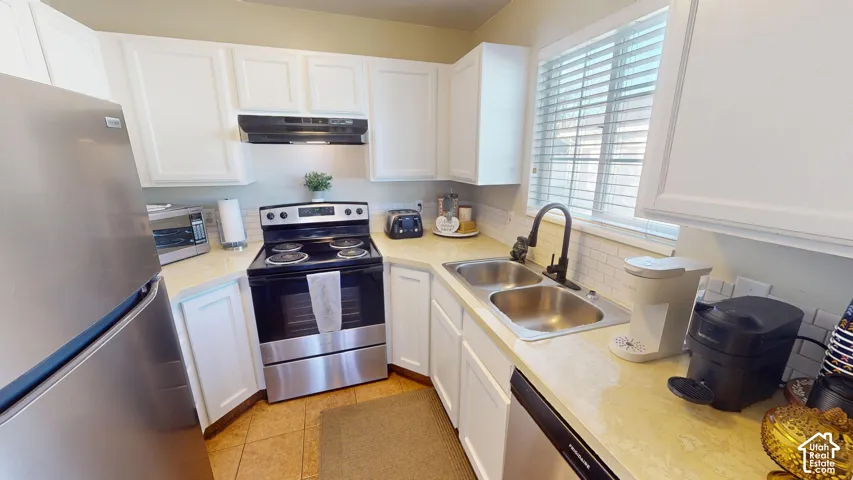 Kitchen featuring white cabinetry, stainless steel appliances, dual-basin stainless steel sink with matte black faucet, tiled backsplash, and solid surface countertops