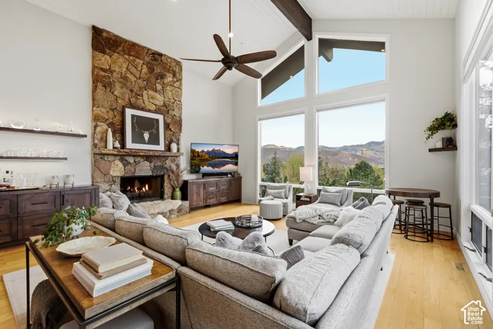 Vaulted ceilings with exposed beams, stone-clad fireplace with hearth, expansive windows, wood-finish flooring, and built-in shelving