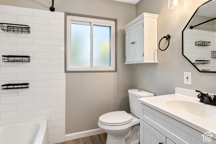 Bathroom featuring white subway tile tub surround, wood-finish flooring, white vanity with integrated basin, wall-mounted storage cabinet, and octagonal mirror with matte black fixtures