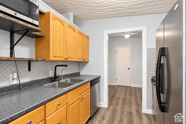 Kitchen featuring wood-finish cabinetry, stone-texture laminate countertops, a hexagonal tile backsplash, stainless steel appliances, and wood-finish flooring