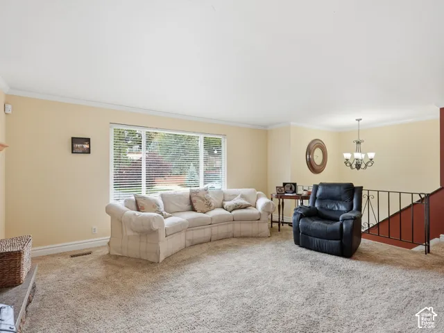 Living area featuring a large picture window, crown molding, wall-to-wall carpeting, a multi-arm chandelier, and decorative metal stair railing