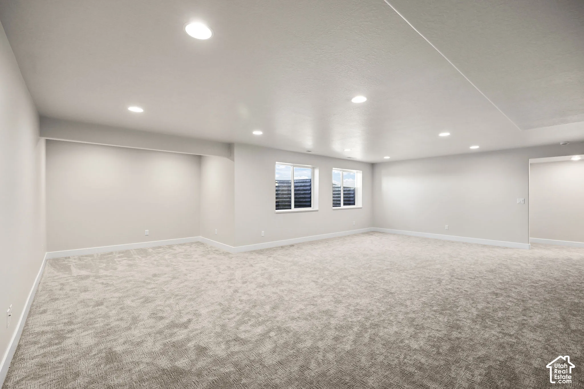 Spacious room featuring light gray carpeting, recessed ceiling lighting, two windows with horizontal blinds, and clean white baseboards