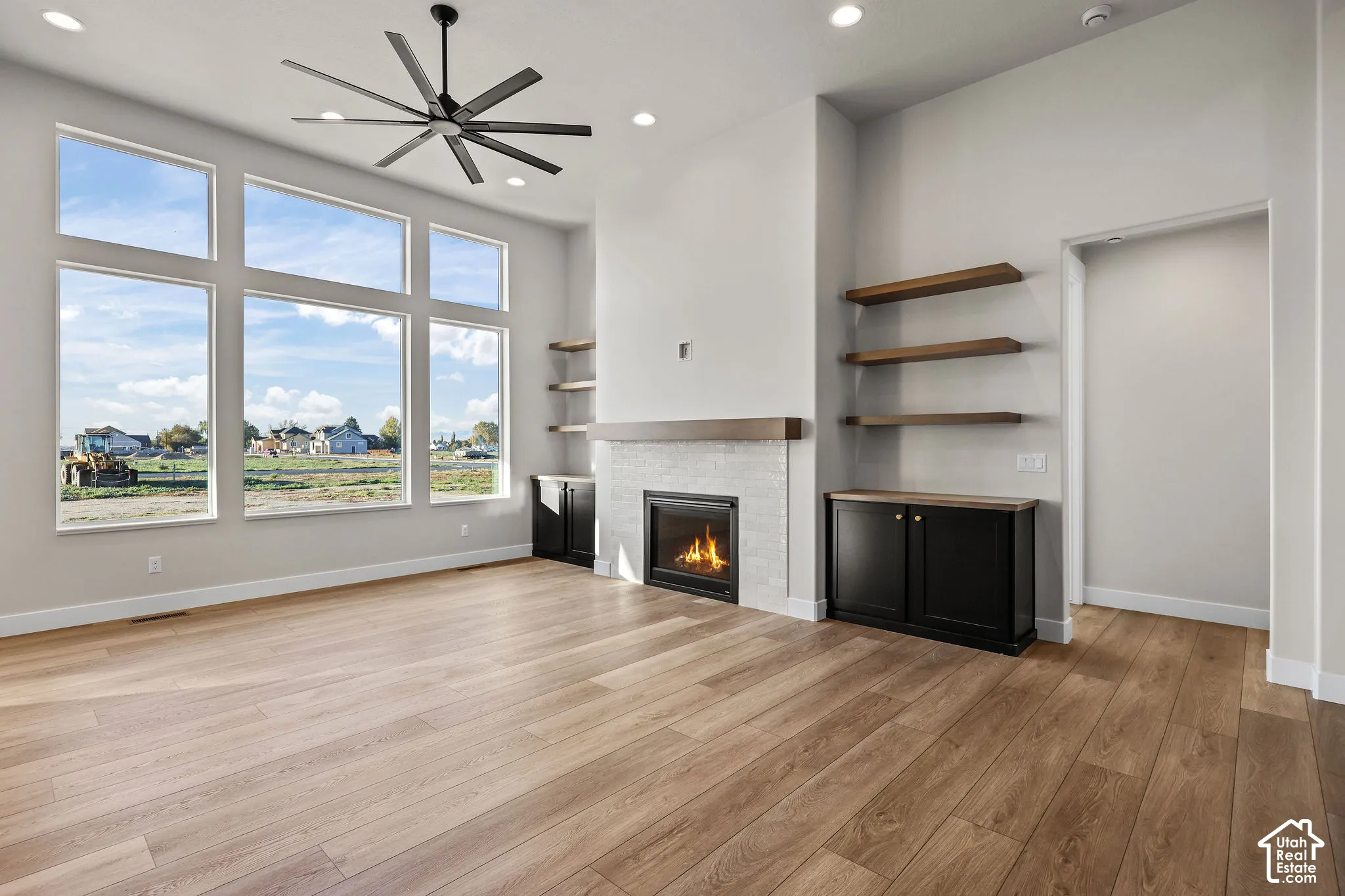Spacious living area featuring expansive windows, wood-finish flooring, a contemporary ceiling fan, and a fireplace with a light-toned surround and wood mantel