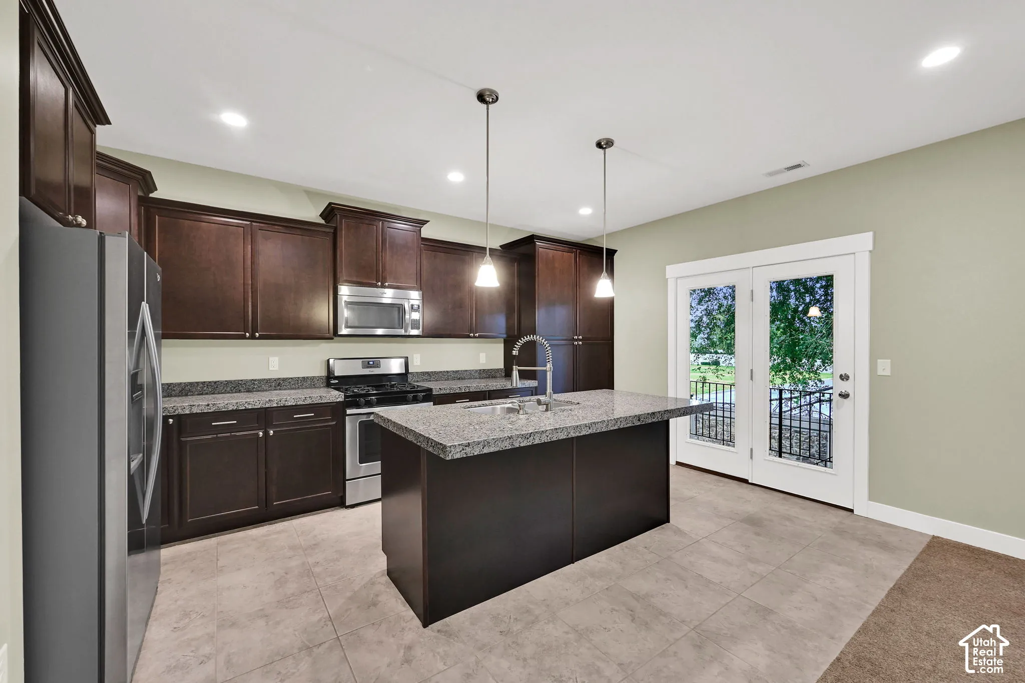 Kitchen featuring dark wood cabinetry, stone countertops, stainless steel appliances, a central island with a built-in sink, and pendant lighting