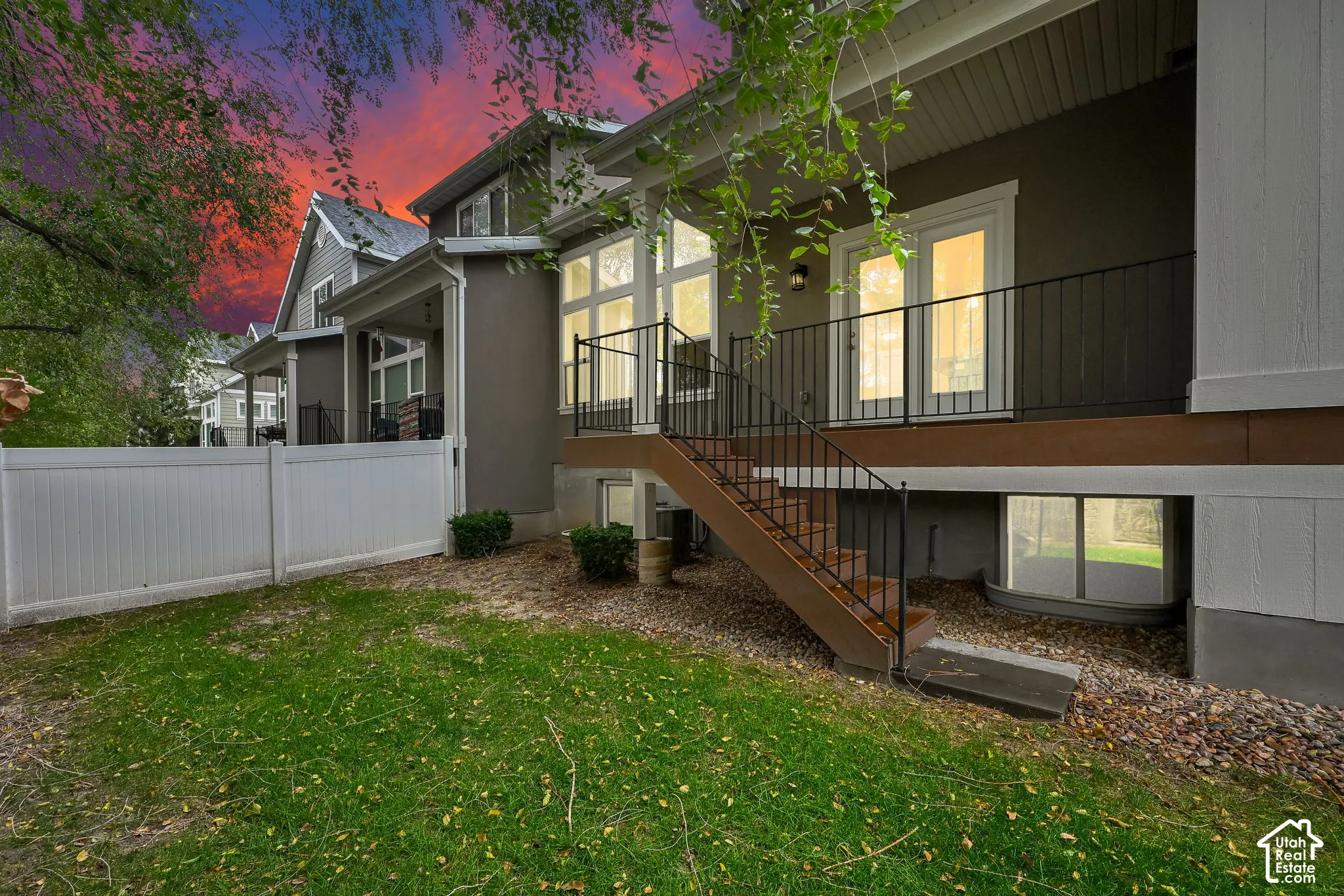 Exterior elevation featuring a raised deck with metal railings, outdoor staircase, white vinyl fencing, and ground-level window well