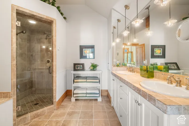 Bathroom featuring a walk-in shower with stone tile surround, dual-sink vanity with stone countertops, full-wall vanity mirror, and pendant lighting
