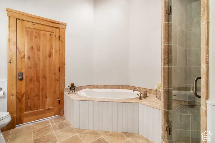 Corner soaking tub featuring a tile surround and beadboard paneling, accompanied by a glass-enclosed shower with stone tile, tile flooring, and natural wood-finish door with trim