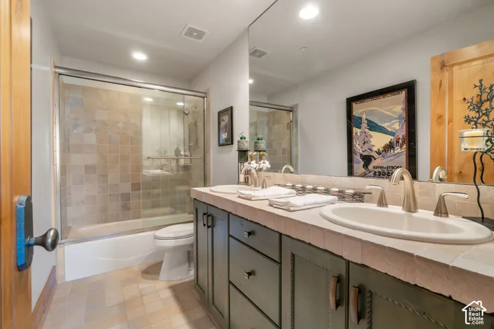 Dual-sink vanity with tile countertop, custom cabinetry, tiled shower enclosure with sliding glass door, and recessed lighting