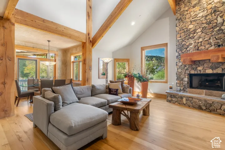 Vaulted ceilings featuring exposed timber beams, full-height stone fireplace with a natural wood mantel, wood-finish flooring, and expansive windows