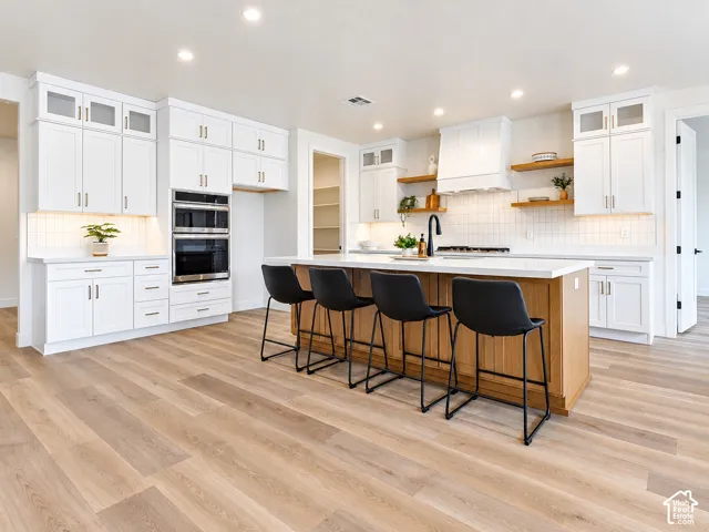 Kitchen featuring white shaker-style cabinetry, wood-finish flooring, central island with quartz countertop, built-in wall oven, and floating wood shelving