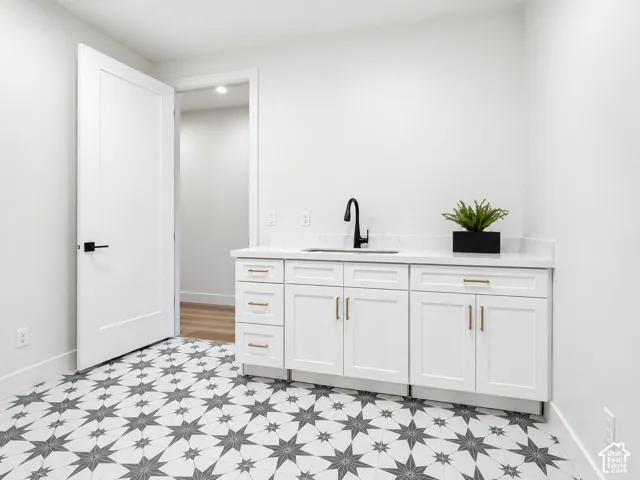 White cabinetry featuring gold-tone hardware, white stone-finish countertop, undermount sink with matte black high-arch faucet, and patterned geometric floor tile