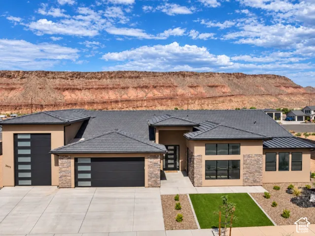 Contemporary single-story exterior featuring multi-textured facade with stone accents, dark-toned tile roof, glass-panel garage doors, and a red rock mesa backdrop