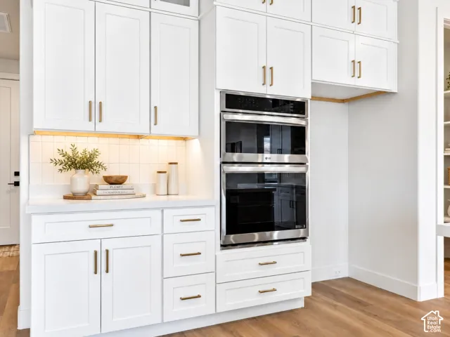 White shaker-style cabinetry with gold-finish hardware, built-in wall ovens, countertop-to-cabinet square tile backsplash, and wood-finish flooring