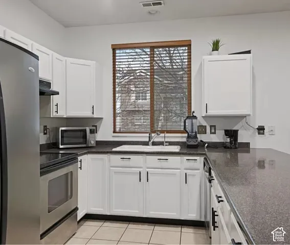 Kitchen featuring white shaker-style cabinetry, stainless steel appliances, dark stone-finish countertops, tile flooring, and a window above the sink
