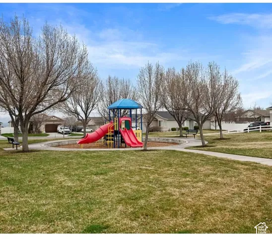 Common outdoor recreation area featuring a playground structure with slides, paved walking paths, benches, and mature trees situated within an expansive lawn