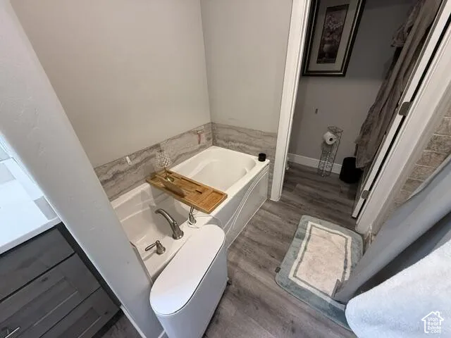Built-in soaking tub with a deck-mounted faucet and neutral stone-finish tile surround, wood-finish flooring, and cabinetry with a white countertop