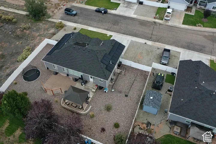 Aerial perspective of a property featuring roof-mounted solar panels, a paved patio with a gazebo, an enclosed backyard with white vinyl fencing, and a gravel landscape