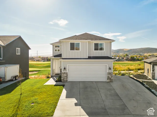 Two-story residence featuring vertical siding, stone veneer accents, an attached two-car garage, and concrete driveway, situated on a lot bordering open terrain