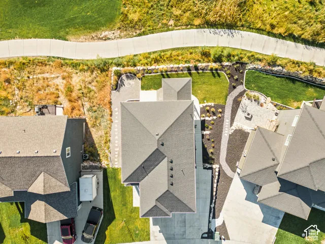 Aerial perspective featuring a multi-pitched shingled roof, concrete driveway, stone-bordered landscaping, and a paver patio