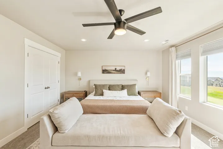 Spacious primary bedroom featuring a ceiling fan with brushed metal finish, abundant natural light from multiple windows with light-filtering treatments, wood-finish flooring, neutral wall finishes, and a substantial sitting area with built-in seating