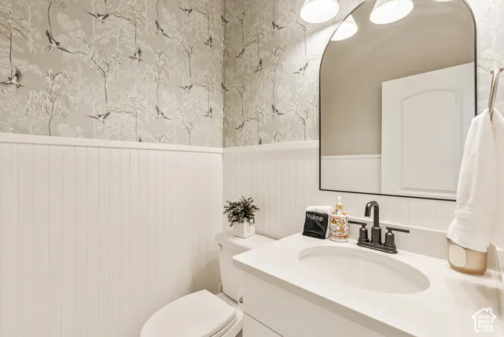 Bathroom featuring beadboard wainscoting, patterned wallpaper, a white vanity with an integrated basin, a matte black faucet, and an arched mirror with a black frame