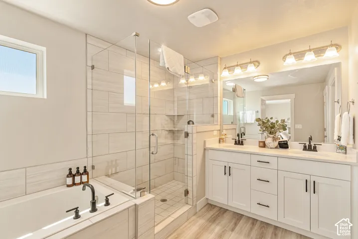 Bathroom featuring a dual-sink vanity with matte black hardware, a tiled walk-in shower with a glass enclosure, a deep soaking tub, and wood-finish flooring