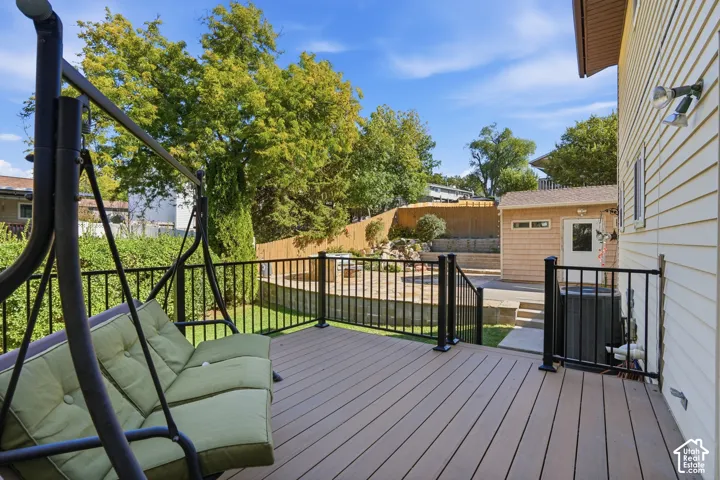 Raised wood-finish deck with black metal railing, multi-level hardscaped patio, stone retaining wall, and detached structure with neutral siding