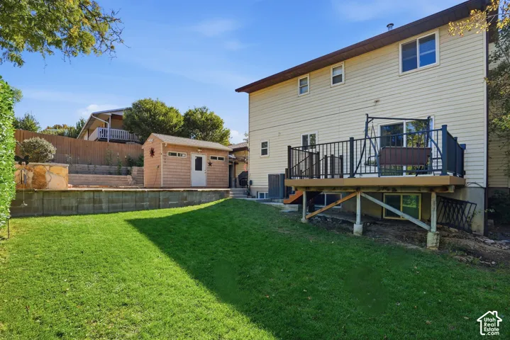 Rear exterior featuring a tiered backyard, elevated composite deck with black metal railing, detached storage structure, and stone retaining walls