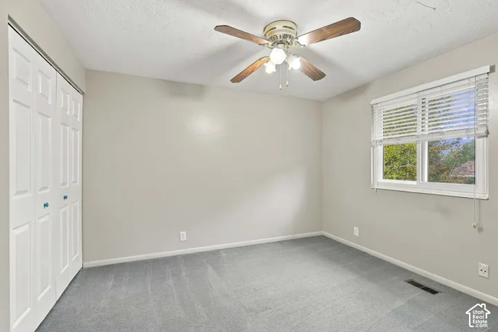 Room with neutral wall color, grey carpeting, ceiling fan with light fixture, sliding closet doors, and a window with horizontal blinds