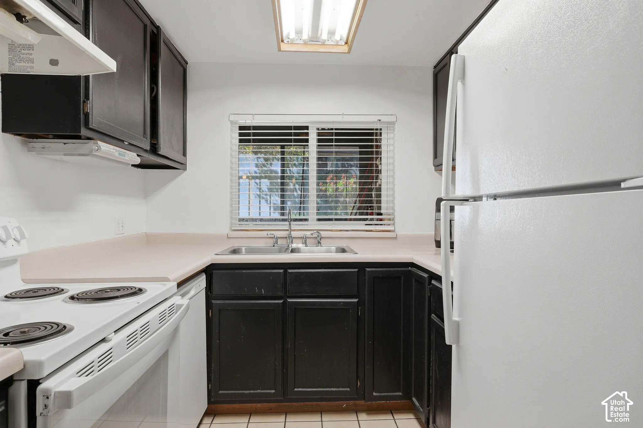 U-shaped kitchen layout featuring dark wood cabinetry, light-toned countertops, a stainless steel sink, and white appliances