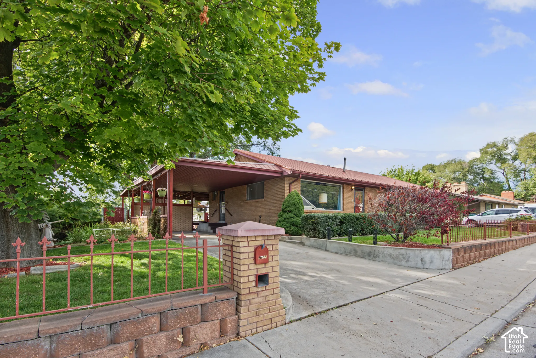 Brick exterior with a covered carport, mature trees, a driveway, and a front yard with a decorative fence