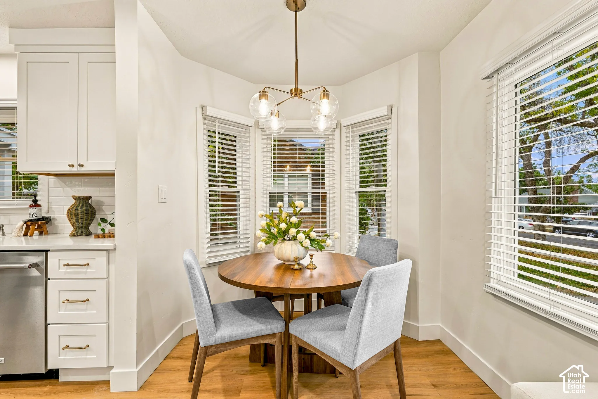 Integrated dining area featuring a bay window with blinds, wood-finish flooring, a contemporary chandelier, and white cabinetry with a subway tile backsplash