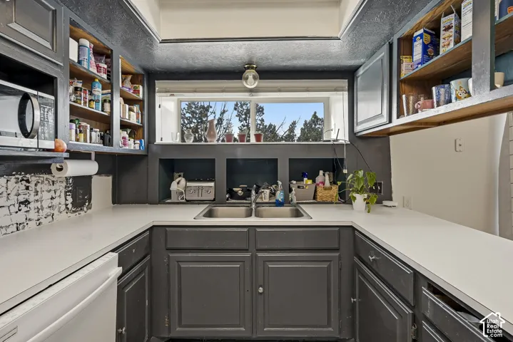 Kitchen featuring charcoal cabinetry, dual-basin stainless steel sink, open upper shelving, solid-surface countertops, and a recessed ceiling light