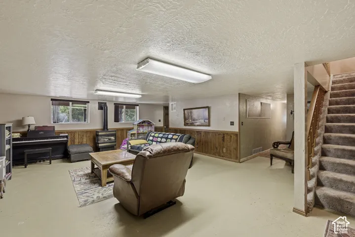 Interior space featuring wood-panel wainscoting, a freestanding stove with a metal flue, dual linear overhead light fixtures, and carpeted staircase with a wood railing