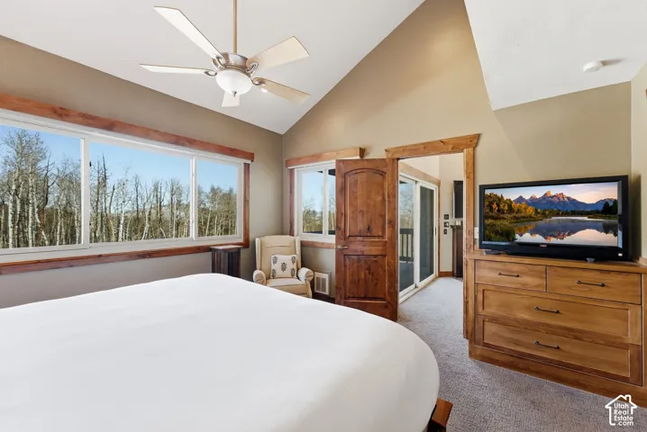 Vaulted ceiling with ceiling fan, large windows with natural scenery, wood trim accents, wood-finish dresser, and carpeted flooring