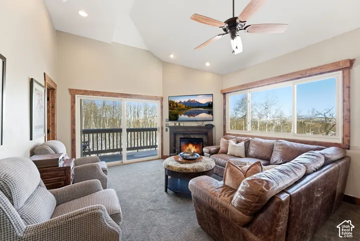 Vaulted ceiling with a central paddle fan, floor-to-ceiling windows, stone-surround fireplace with a wood mantel, and sliding glass door access to an exterior deck