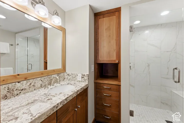 Spa-inspired master bathroom featuring a dual-sink vanity with speckled stone countertop, wood-framed mirror with integrated lighting, custom wood cabinetry with multiple drawers, walk-in shower with marble-look tile surround and frameless glass enclosure