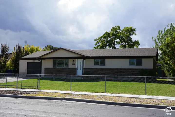 Single-story residence featuring a modern exterior with contrasting brick and siding, attached garage, front-facing entry, manicured lawn, mature landscaping, and chain-link perimeter fencing
