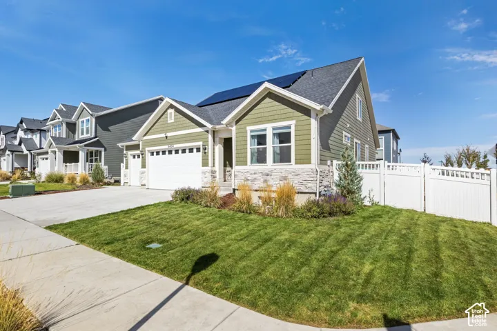 Exterior featuring olive-toned horizontal siding, a stone veneer base, an attached two-car garage, roof-mounted solar panels, and white vinyl perimeter fencing