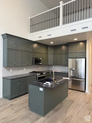 Kitchen featuring dark sage cabinetry, stone-surface countertops, herringbone-pattern tile backsplash, stainless steel appliances, and a center island with an undermount sink and metallic faucet, beneath a mezzanine level with black wrought-iron railings