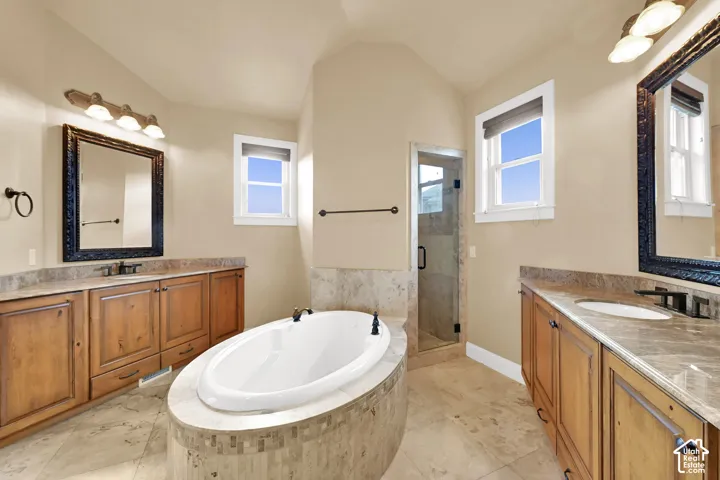 Dual wood-finish vanities with stone countertops, a central soaking tub with stone surround, a tiled walk-in shower with glass door, and large rectangular mirrors